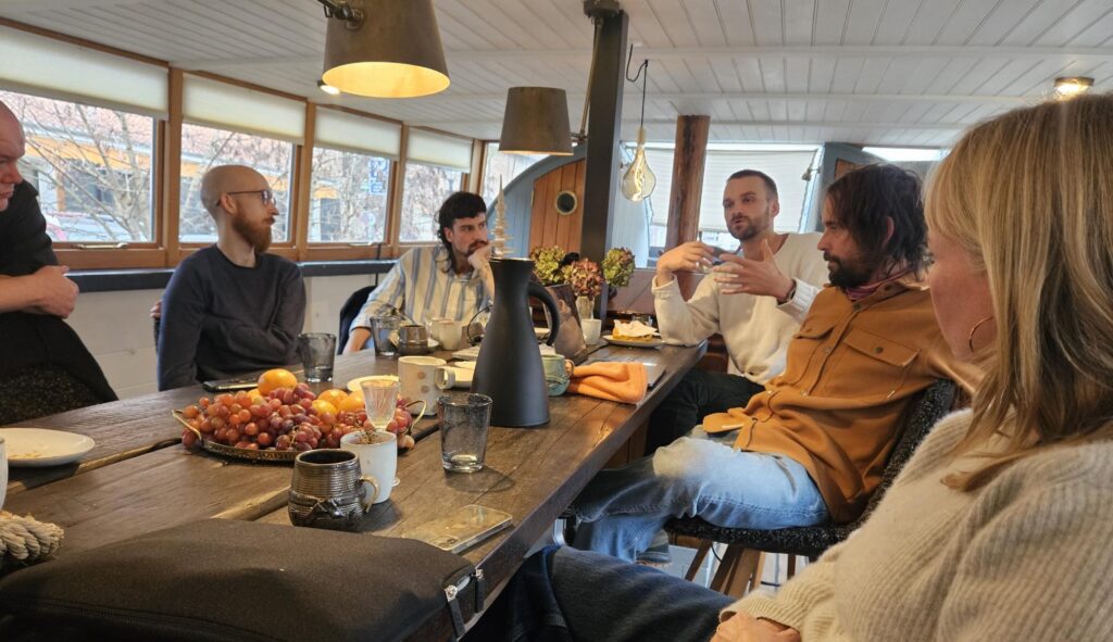 Six people sitting around table made of wood. Five males and one female. There are grapes, glasses and cups on the table.