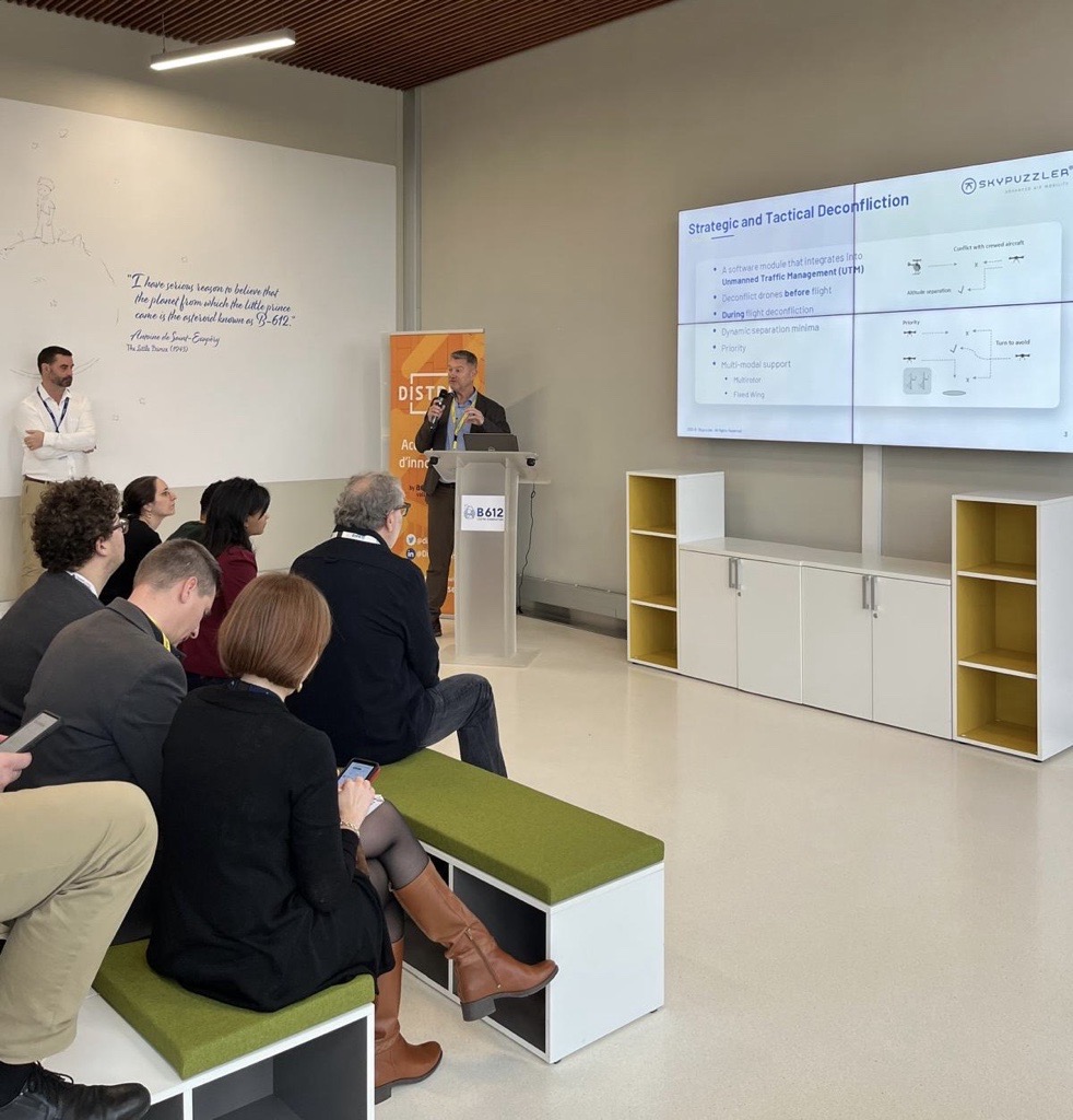 Male standing in front of laptop in front of crowd with a screen behind him showing his presentation