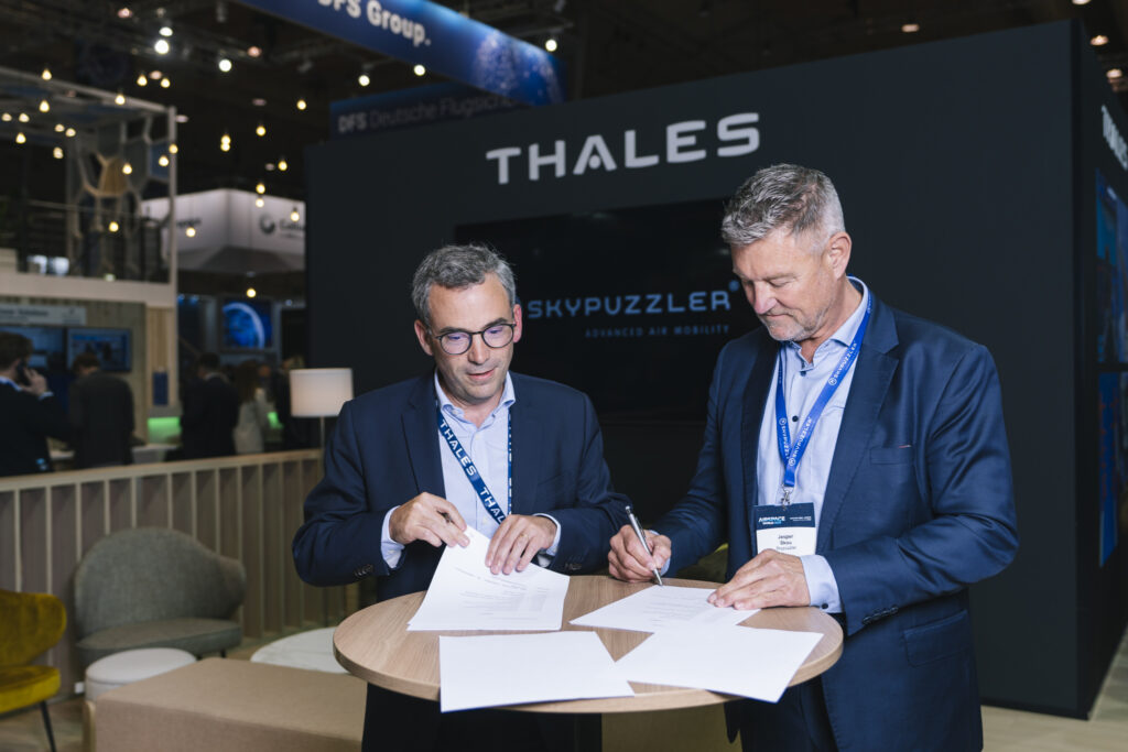 Two males with grey hair and blue suits standing next to each other and signing papers.