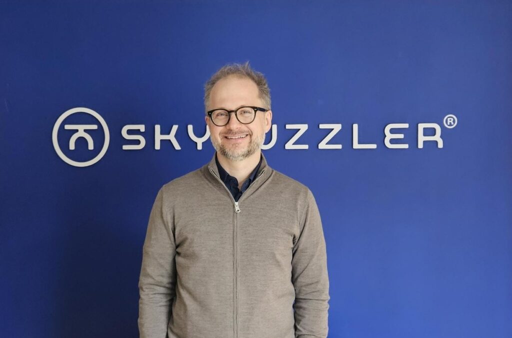 Male with grey sweatshirt and glasses standing in front of blue wall and smiling