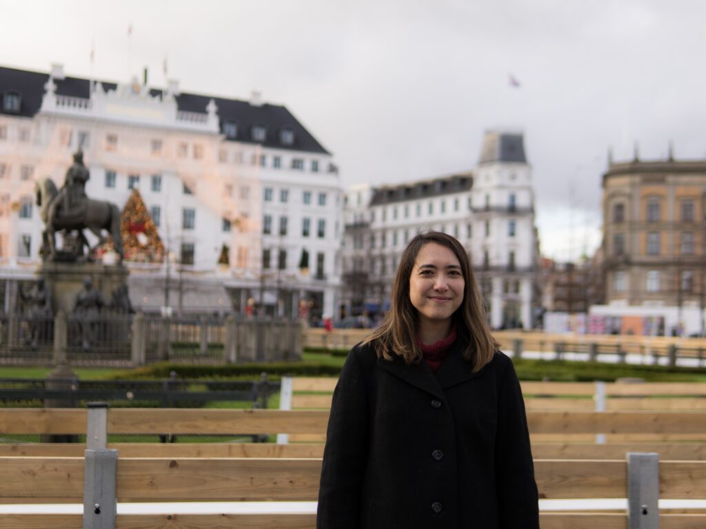 Female with brown hair and black jacking standing in front of a white building with black roof and a christmas tree